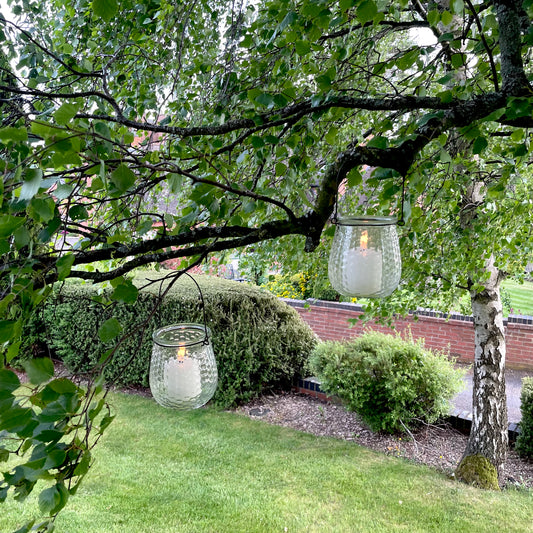 Clear glass hanging candle holder with metal handle, styled outdoors with a glowing tea light.