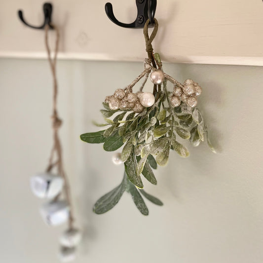 Glittered mistletoe sprig with white berries hanging from a rustic hook against a festive background.