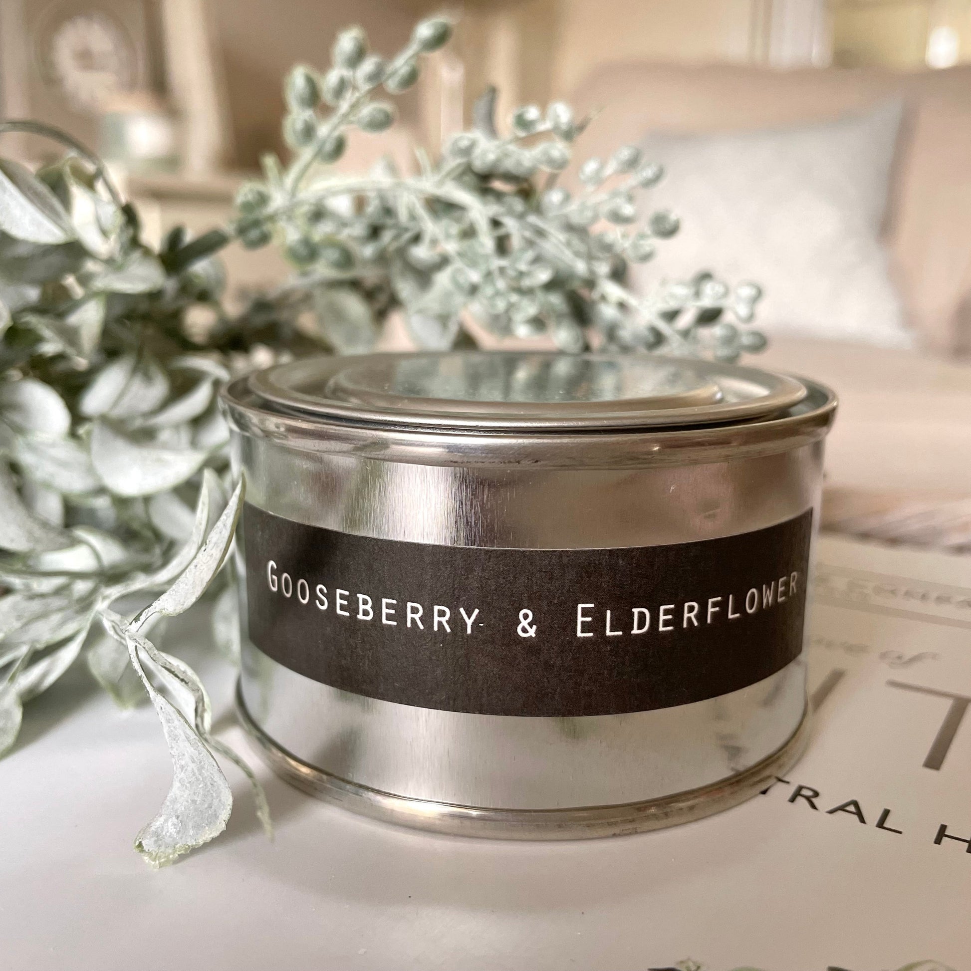Candle labeled 'Gooseberry & Elderflower' on a surface with greenery in the background