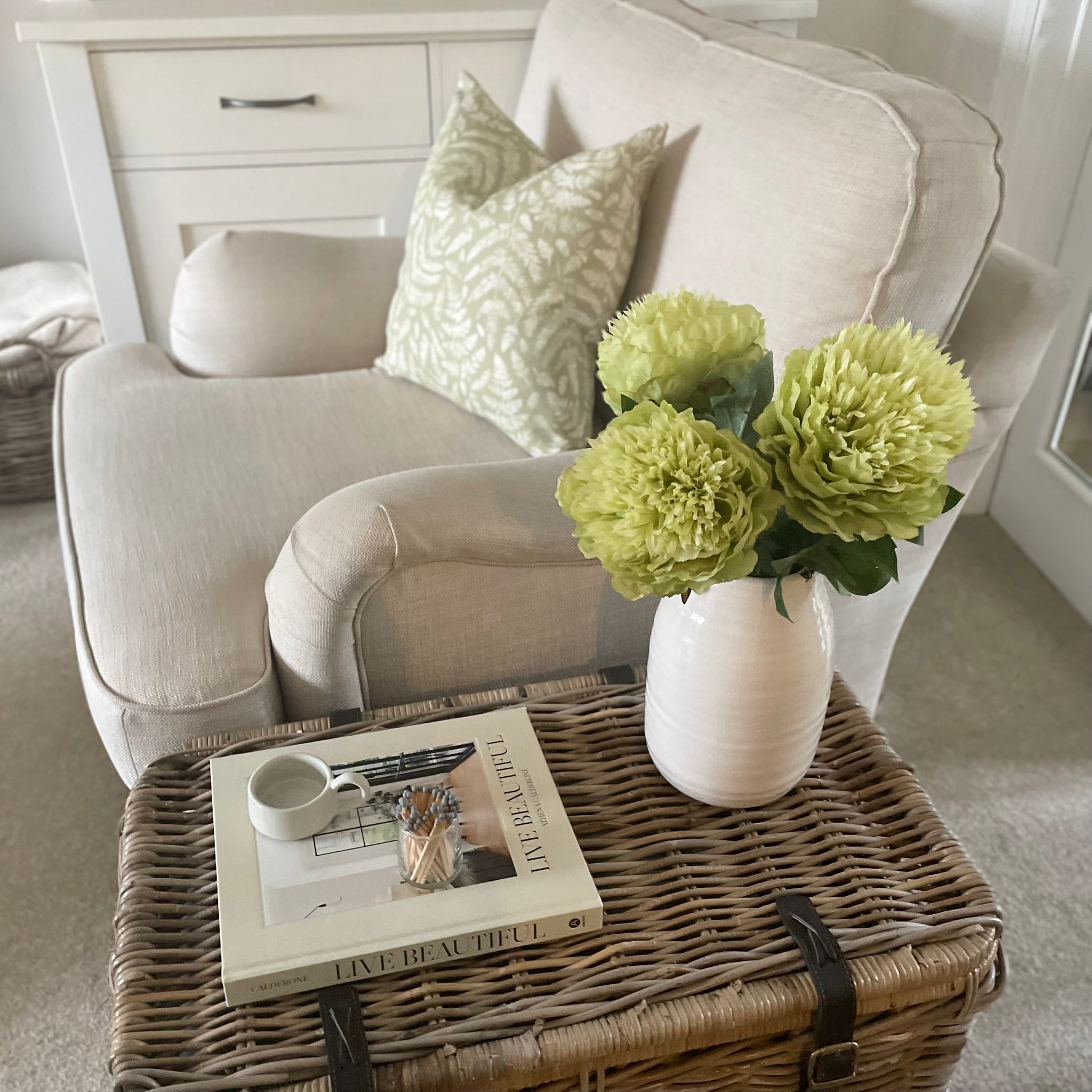 Faux garden peony stem with bright green bloom and leaves. Styled on wicker trunk next to a cream sofa.