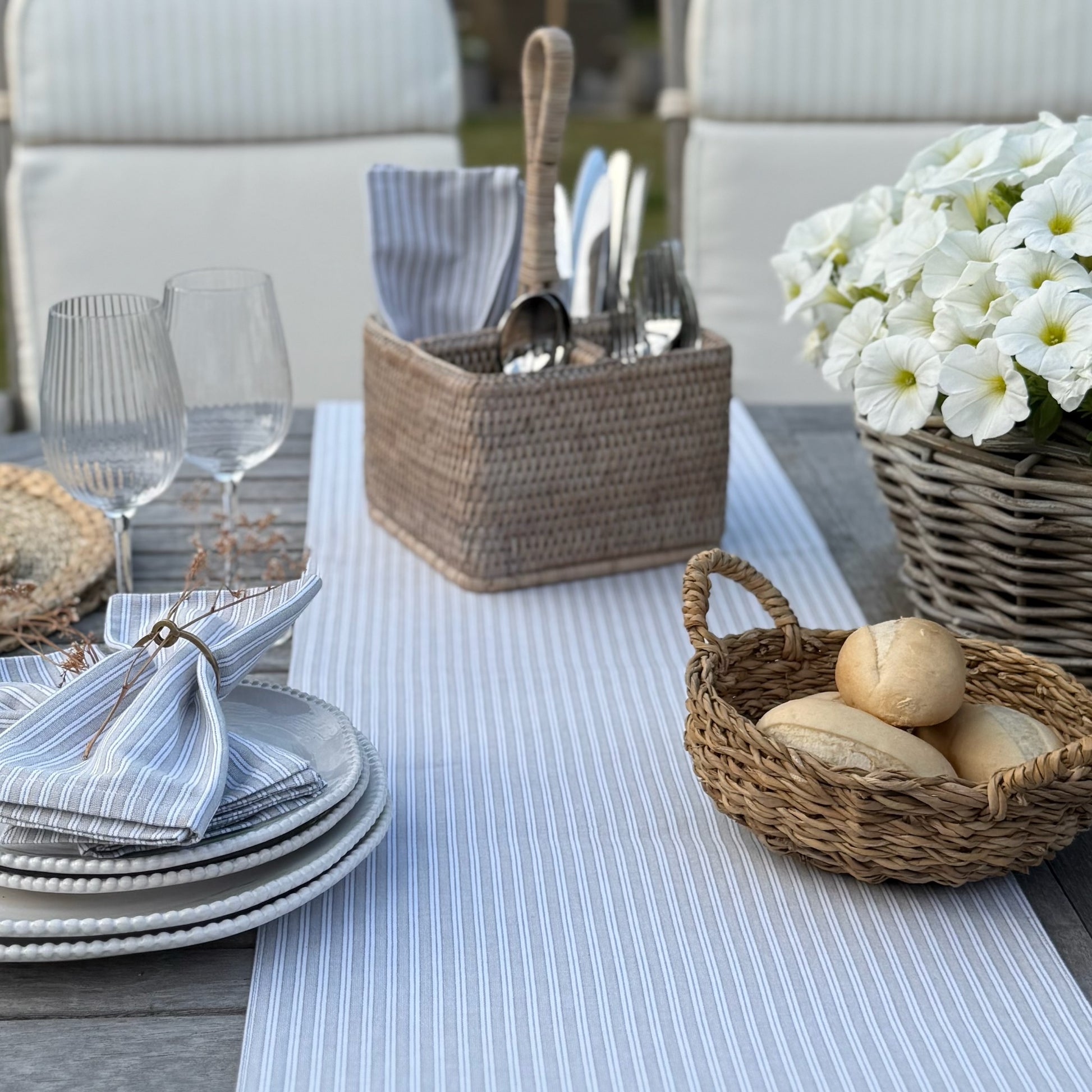Outdoor dining table setting with bread basket, flowers, and cutlery on a striped grey and white table runner.