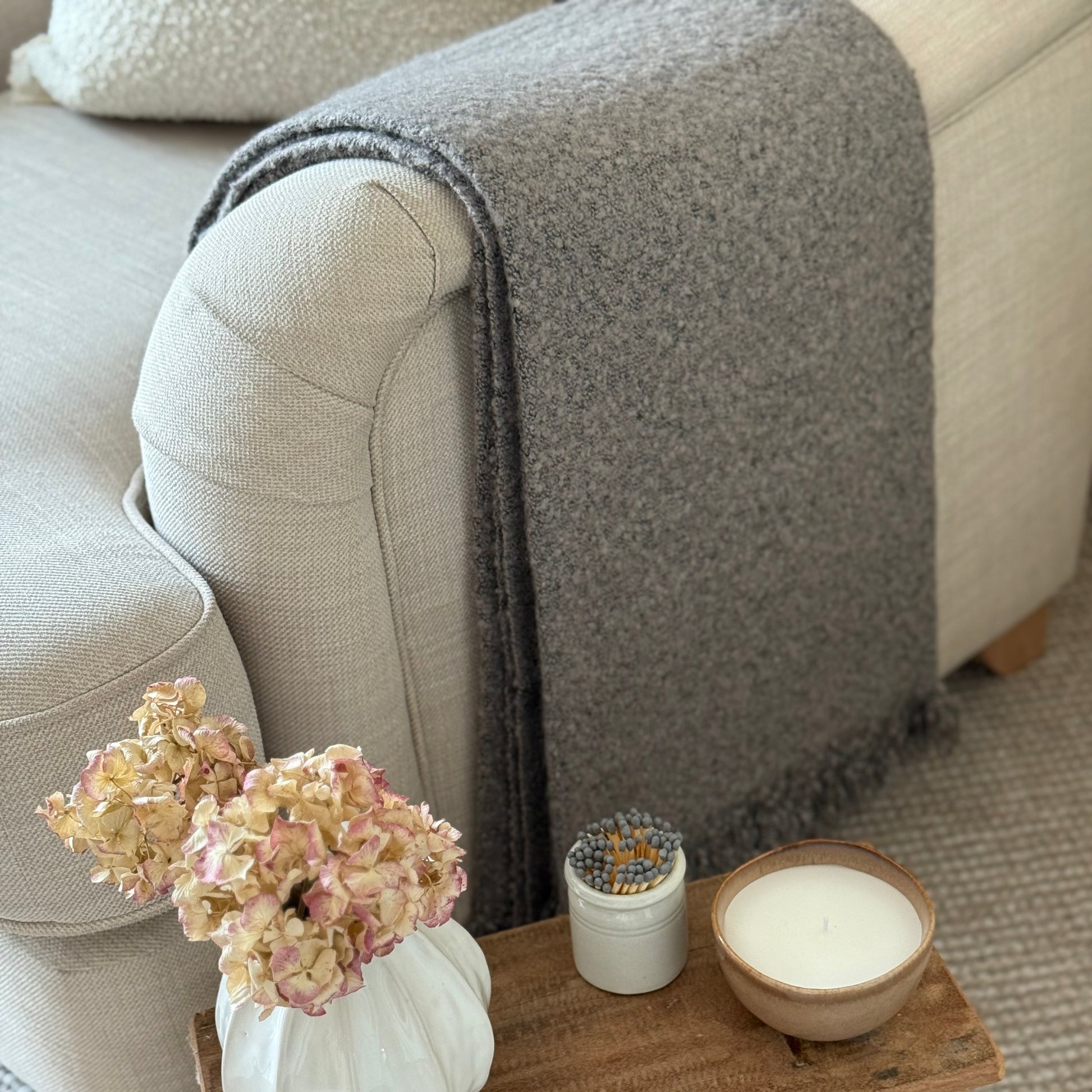 Decorative items including a vase with flowers, small plant, and candle on a wooden tray in front of a gray blanket draped over a sofa.