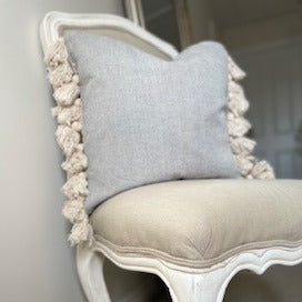 Grey textured cotton cushion with cream tassels on each edge, styled on a neutral chair in a cosy living room setting.