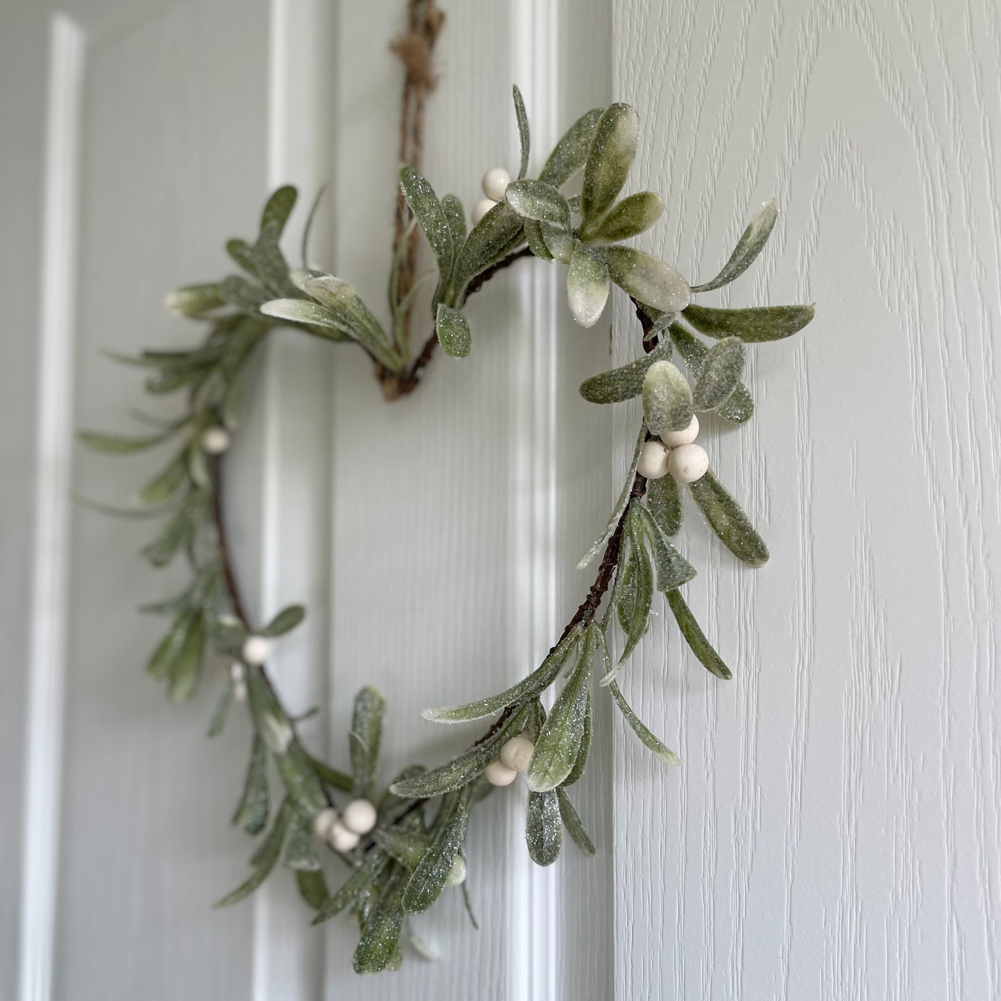 Heart shaped glittered mistletoe wreath with white berries, styled on a white door for a festive touch.