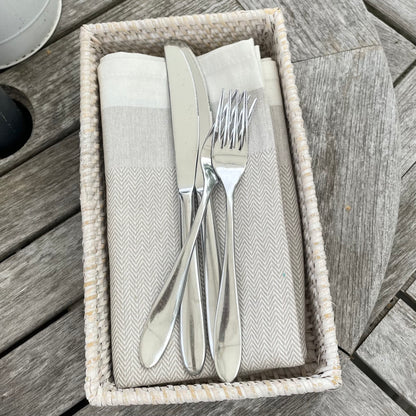 Machine washable taupe herringbone cotton napkin styled with tableware in a rattan rectangular tray.