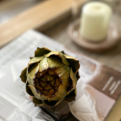 Natural looking artificial artichoke styled on a book with a candle on a wooden coffee table.