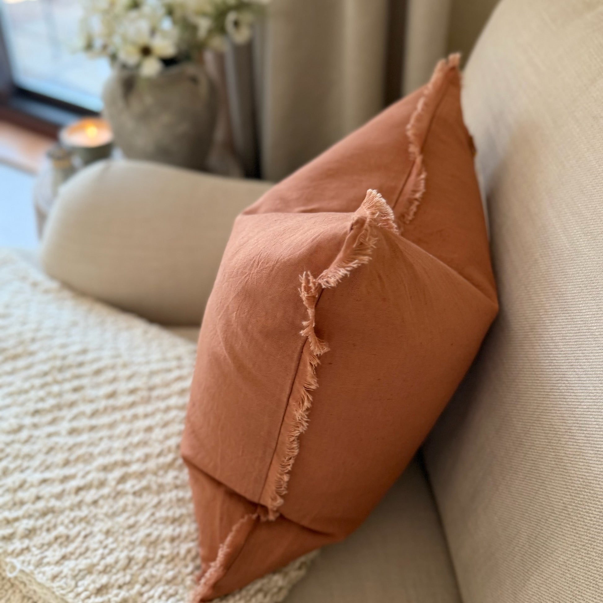 Rust coloured cushion with frayed edges on a beige couch in a cosy living room.