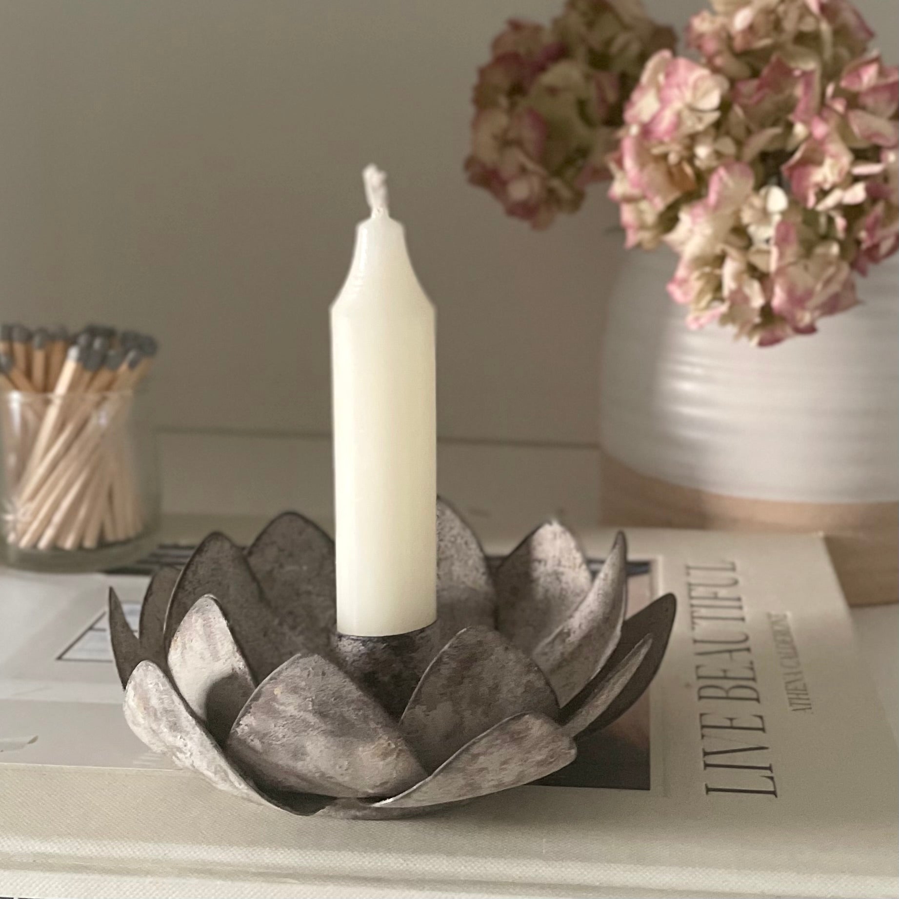Rustic lotus shaped candle holder in zinc metal. Styled with a candle on a book with a pot of flowers in the background.