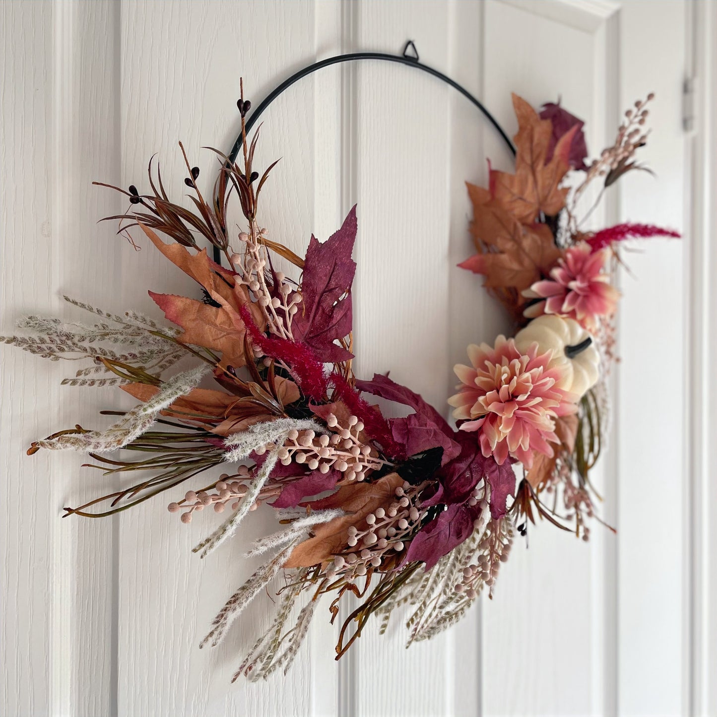 Autumn hoop wreath with pampas grasses, maple leaves, pink berries, pink flowers and a neutral pumpkin. Styled on white door.