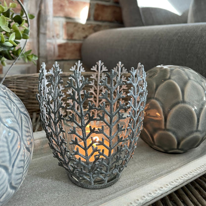 Metal leaf effect tea light holder with glass insert, styled on a neutral wooden surface with a lit candle inside.