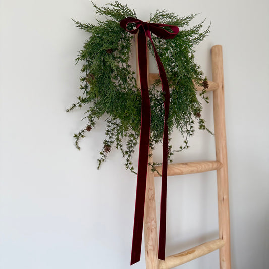 Wooden ladder with a green mini pinecone wreath and red ribbon against a white wall.