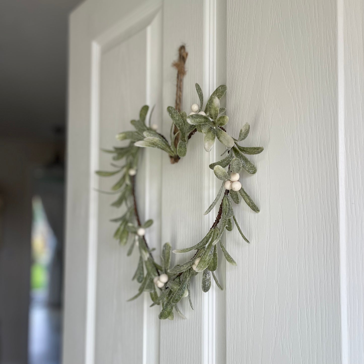 Heart shaped glittered mistletoe wreath with white berries, styled on a white door for a festive touch.