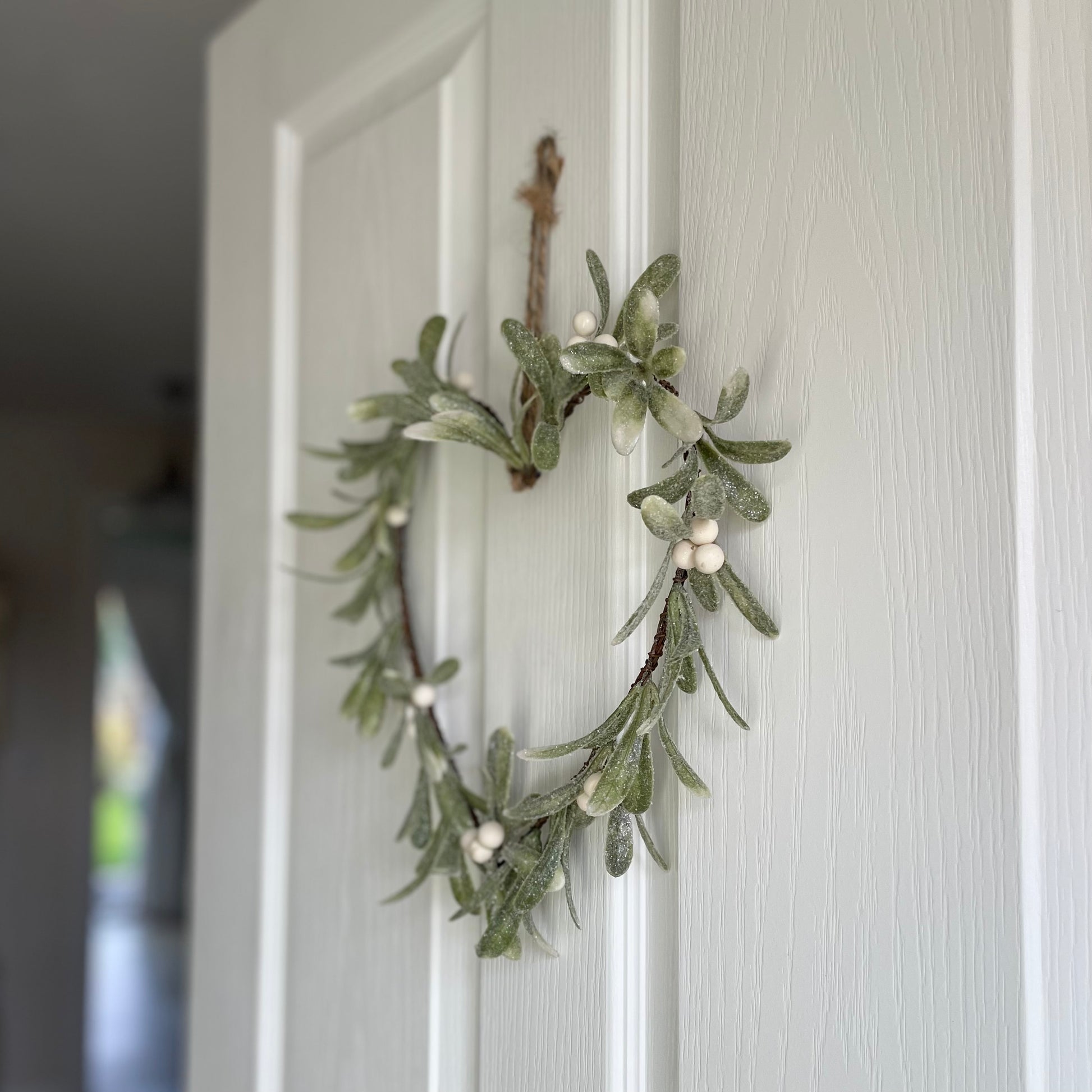 Heart shaped glittered mistletoe wreath with white berries, styled on a white door for a festive touch.