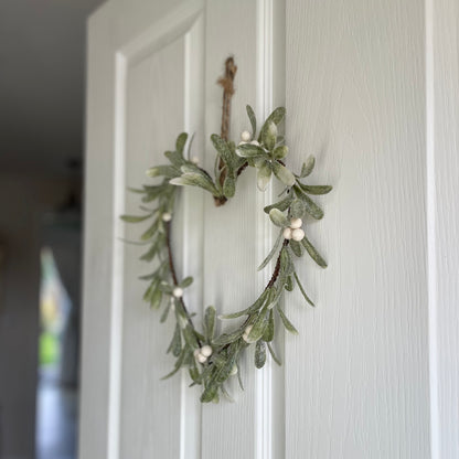 Heart shaped glittered mistletoe wreath with white berries, styled on a white door for a festive touch.