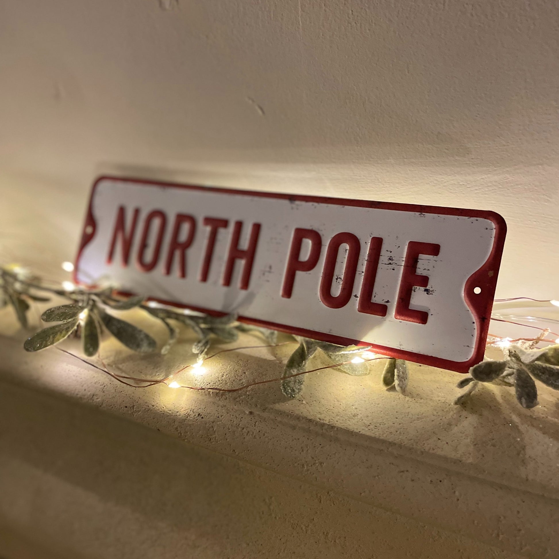 North pole metal sign styled on a mantle piece with fairy lights and foliage.