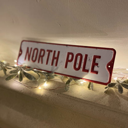 North pole metal sign styled on a mantle piece with fairy lights and foliage.