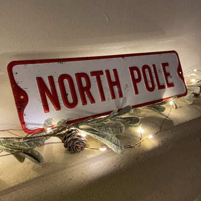North pole metal sign styled on a mantle piece with fairy lights and foliage.