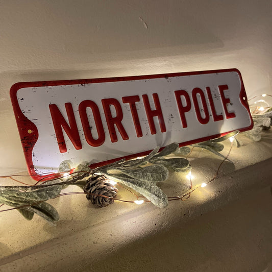 North pole metal sign styled on a mantle piece with fairy lights and foliage.