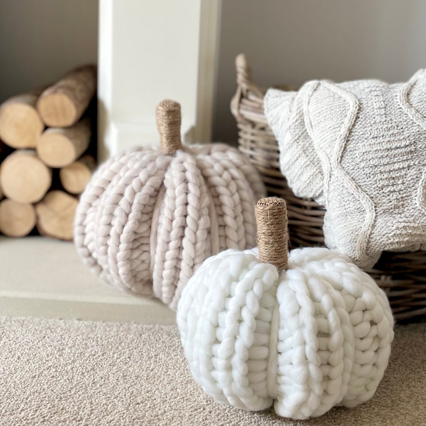 Oatmeal chunky knitted pumpkin with twine-wrapped stalk styled next to a fireplace in a cosy living room.