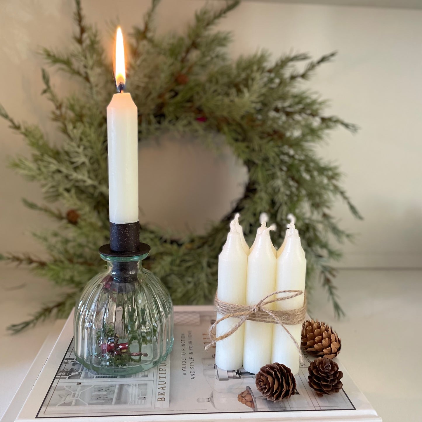 Bundle of six short Danish style dinner candles tied with jute cord, styled on a book with candle and pinecone wreath.