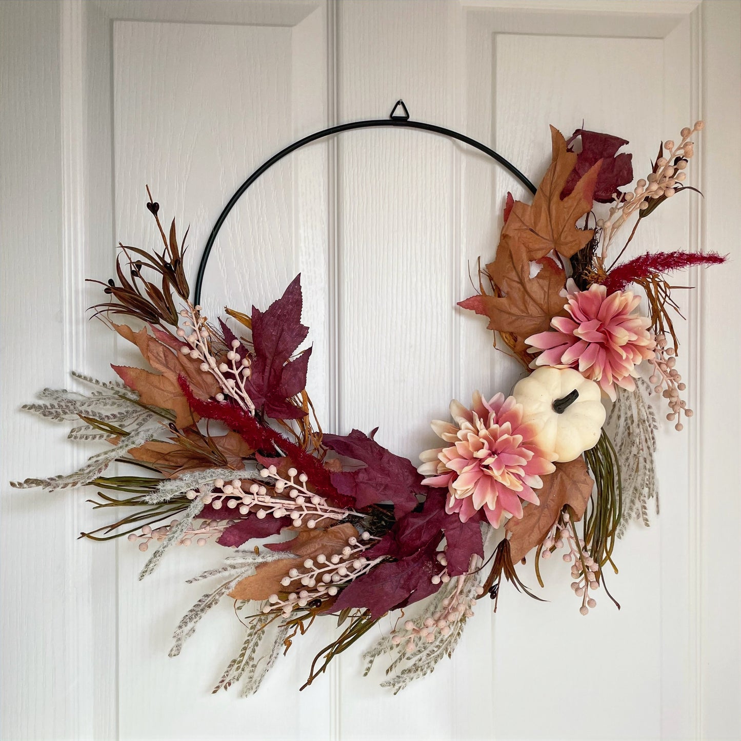 Autumn hoop wreath with pampas grasses, maple leaves, pink berries, pink flowers and a neutral pumpkin. Styled on a white door.