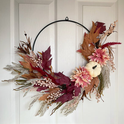 Autumn hoop wreath with pampas grasses, maple leaves, pink berries, pink flowers and a neutral pumpkin. Styled on a white door.