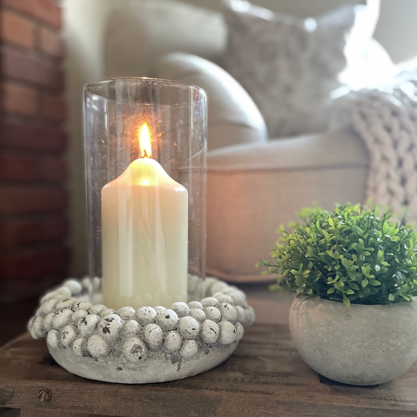 Glass hurricane lantern with aged pebble base. Styled on a rustic wooden stool with a hebe faux plant.