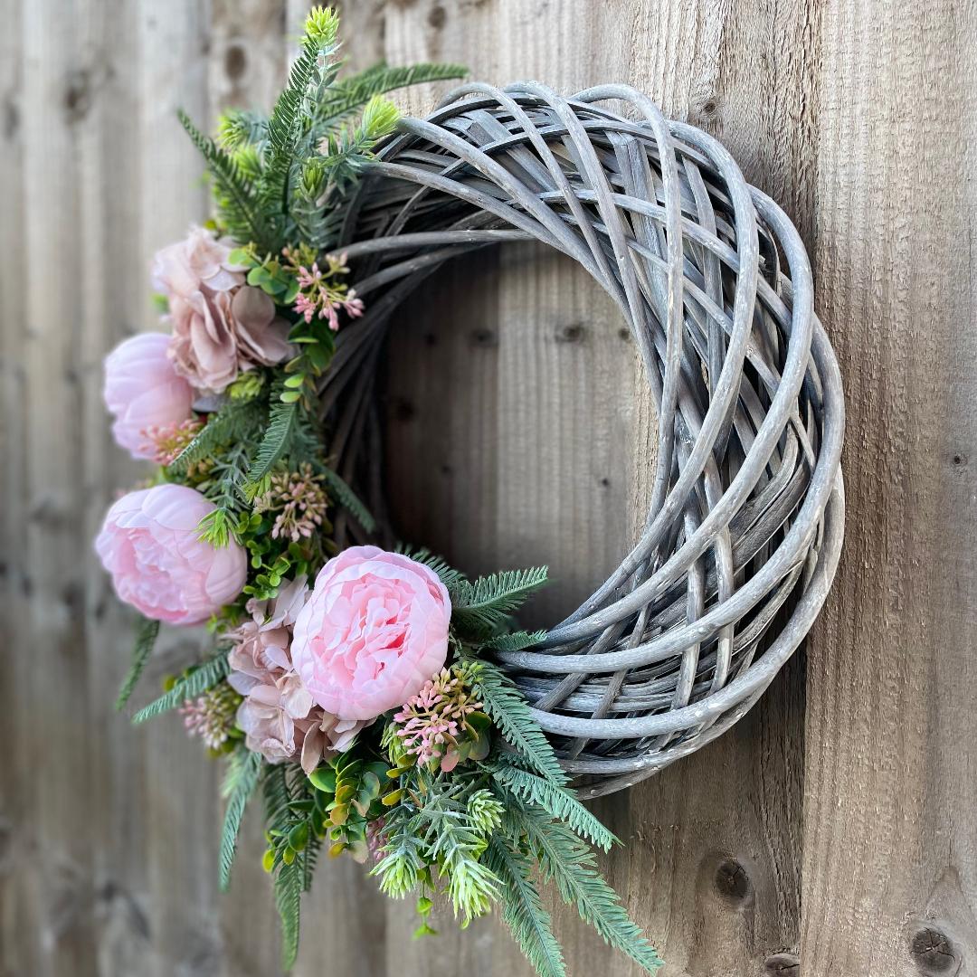 Spring summer wreath with pink peonies and hydrangeas on rattan grey base, styled on a wooden garden fence.