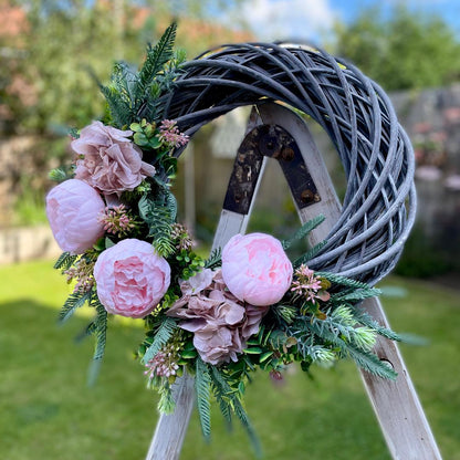 Pink peony and nude hydrangea wreath presented on a wooden ladder. Shown in a garden.