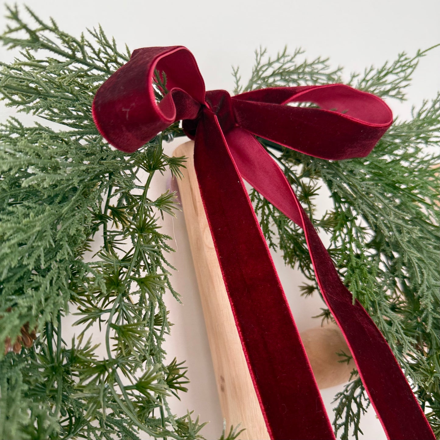 Red ribbon tied on a mini pinecone wreath on a wooden ladder shown on a white background.