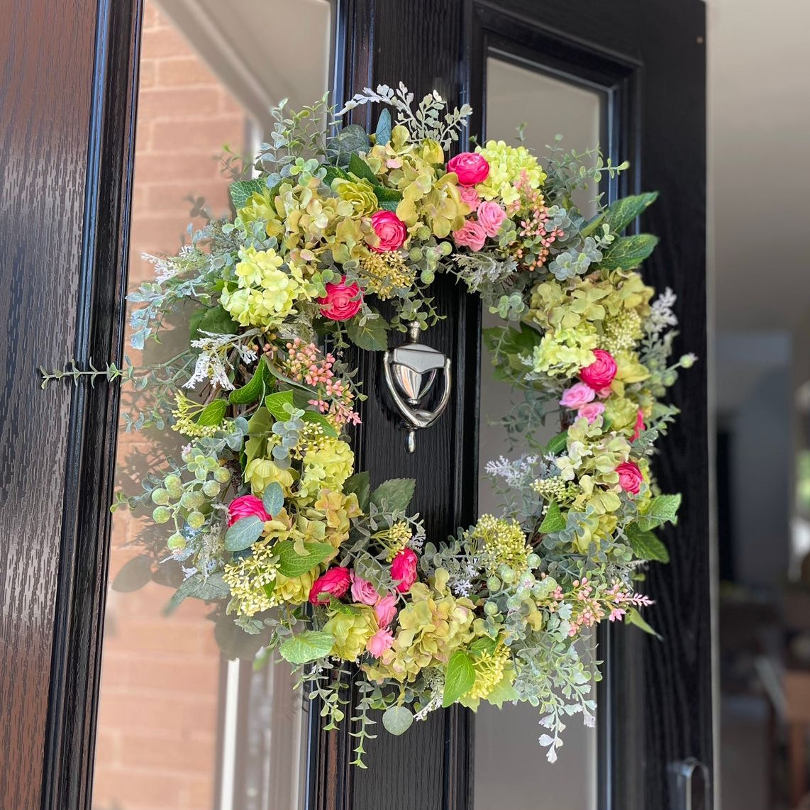 Floral wreath with green and pink flowers on a black front door. Handmade summer wreath with hydrangeas, roses and ranunculus on grapevine base.