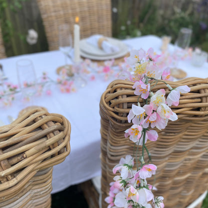 Pink cherry blossom garland with delicate lifelike petals and hanging loops. Draped over a dining table chair in a rose garden.