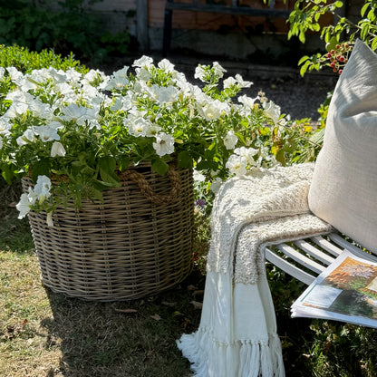White flowers in a woven basket next to a white bench with a blanket and magazine in a garden setting.