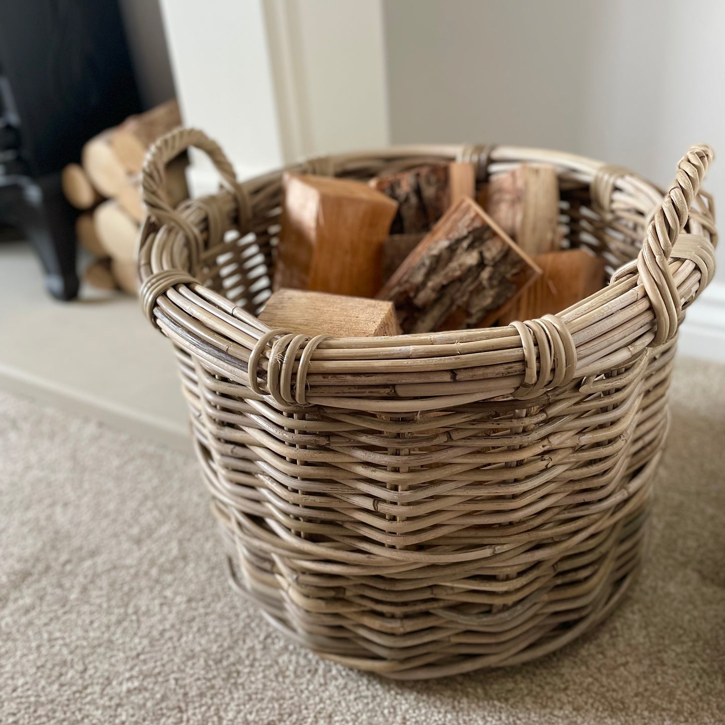 Round rattan basket holding logs by a fireplace in a neutral, cosy interior.