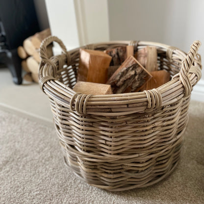 Round rattan basket holding logs by a fireplace in a neutral, cosy interior.