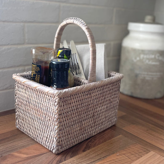 Whitewashed rattan caddy with handle for condiments and napkins styled on a wooden kitchen worktop.