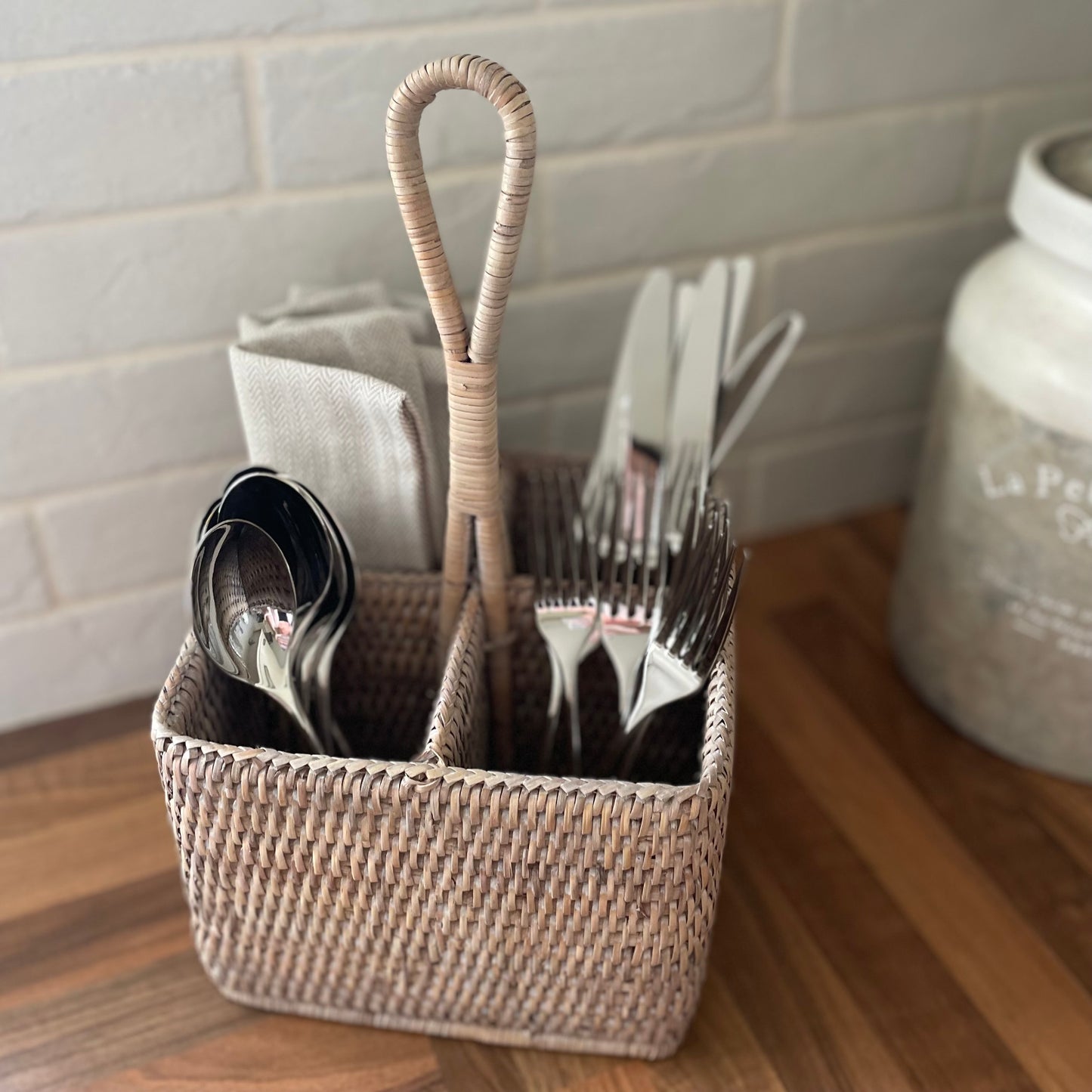Whitewashed rattan caddy with handle for cutlery or condiments. Shown styled on a wooden worktop.
