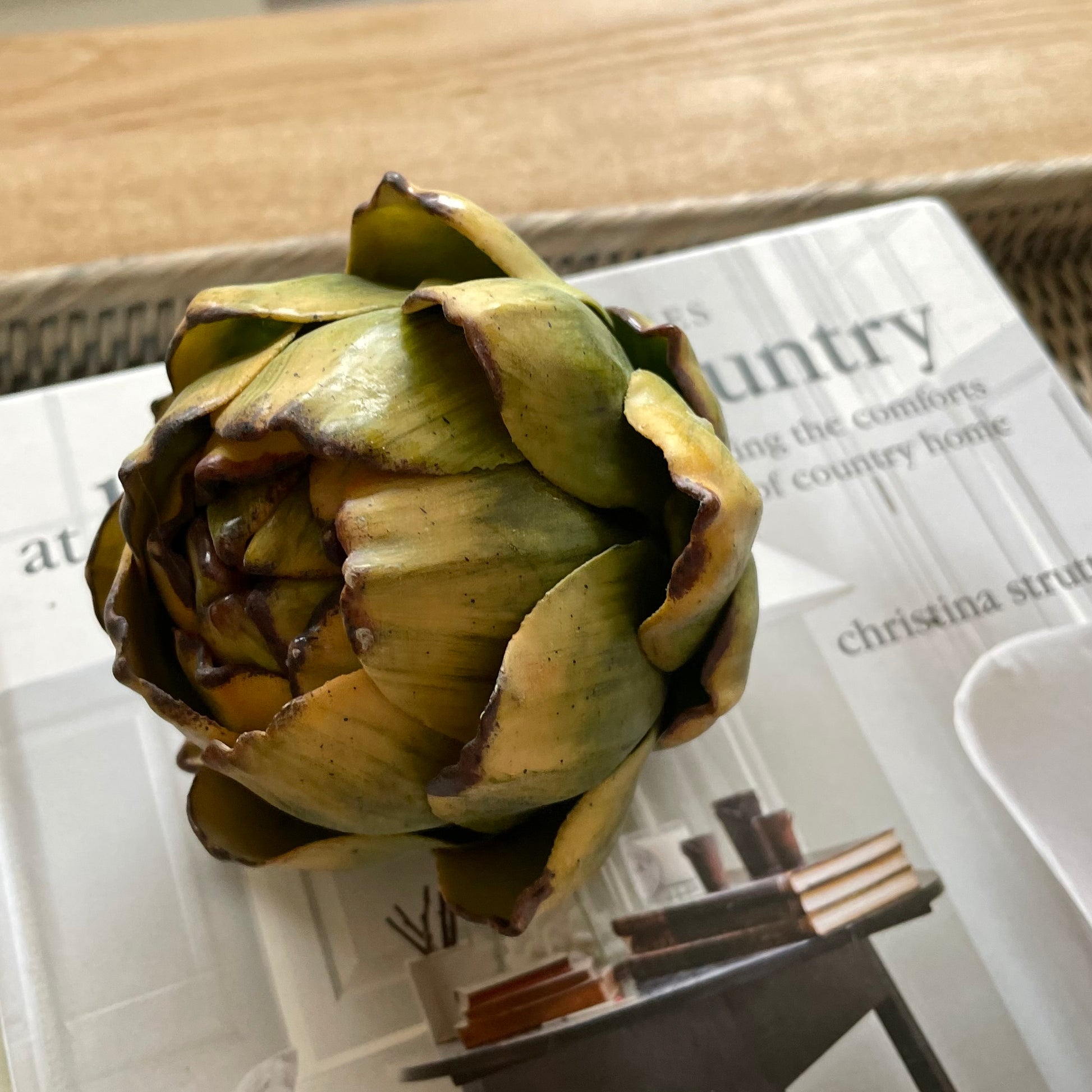 Lifelike faux green artichoke styled on a book on a wooden coffee table.