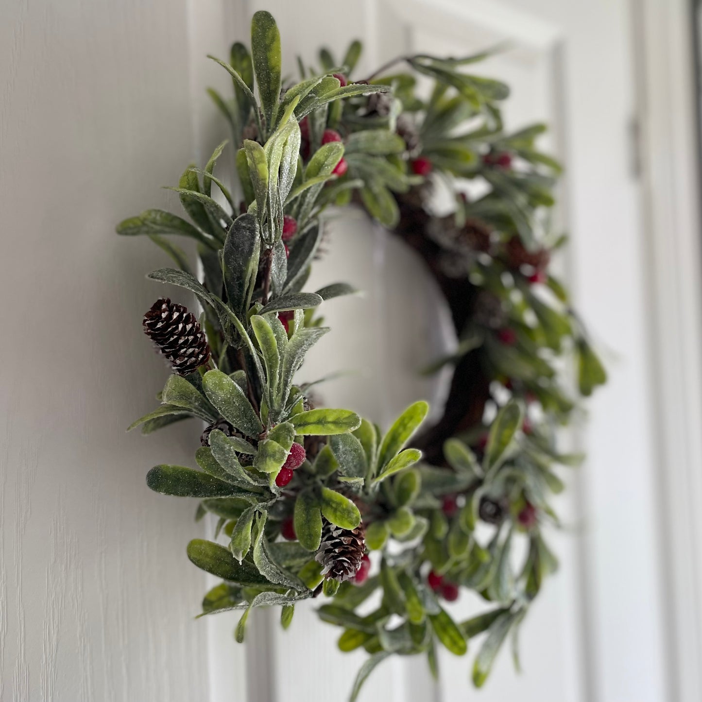 Frosted faux Christmas wreath with green foliage, red berries and fir cones, styled on a white door.