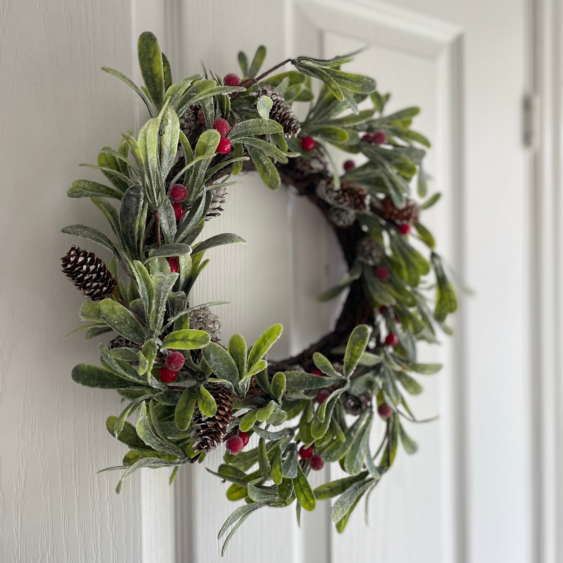 Frosted faux Christmas wreath with green foliage, red berries and fir cones, styled on a white door.