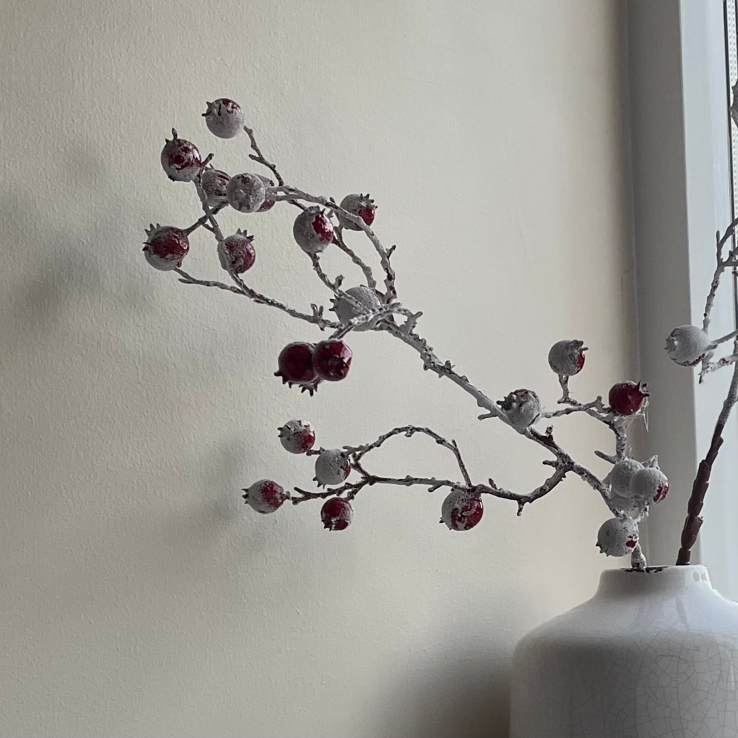 Close up of realistic faux red berries on brown stem, styled in a white crackled ceramic vase.