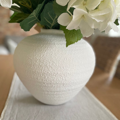 White matte roman urn style Regola vase with soft textured finish and elegant curved shape. Styled on a dining table with a white hydrangea bouquet.