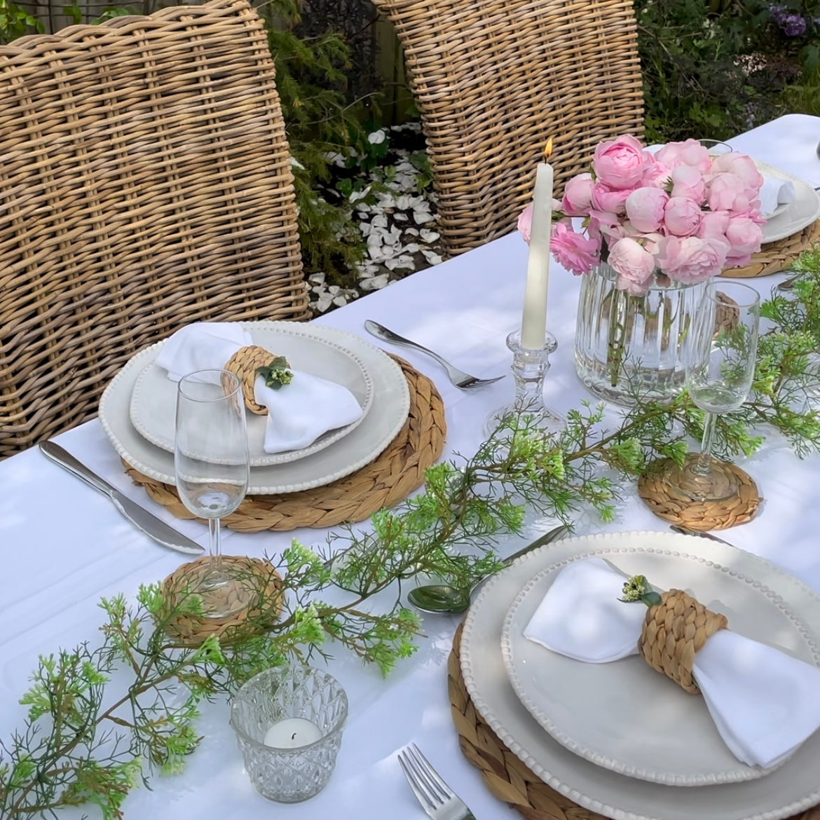 Set of round woven hyacinth coasters arranged on a table, showing their natural texture and warm, organic colour. Styled in a garden with roses.