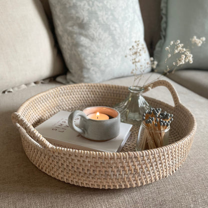 White washed round rattan tray with handles styled on a sofa with home accessories.