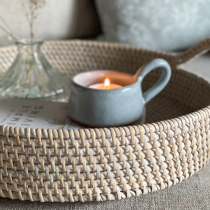 Whitewashed round rattan tray with handles styled on a settee with home accessories.