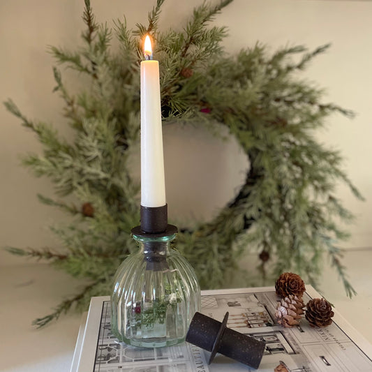 Close up of bottle top candle holder on a glass bud vase. Styled on a book with a wreath in the background. Candle lit for ambience.