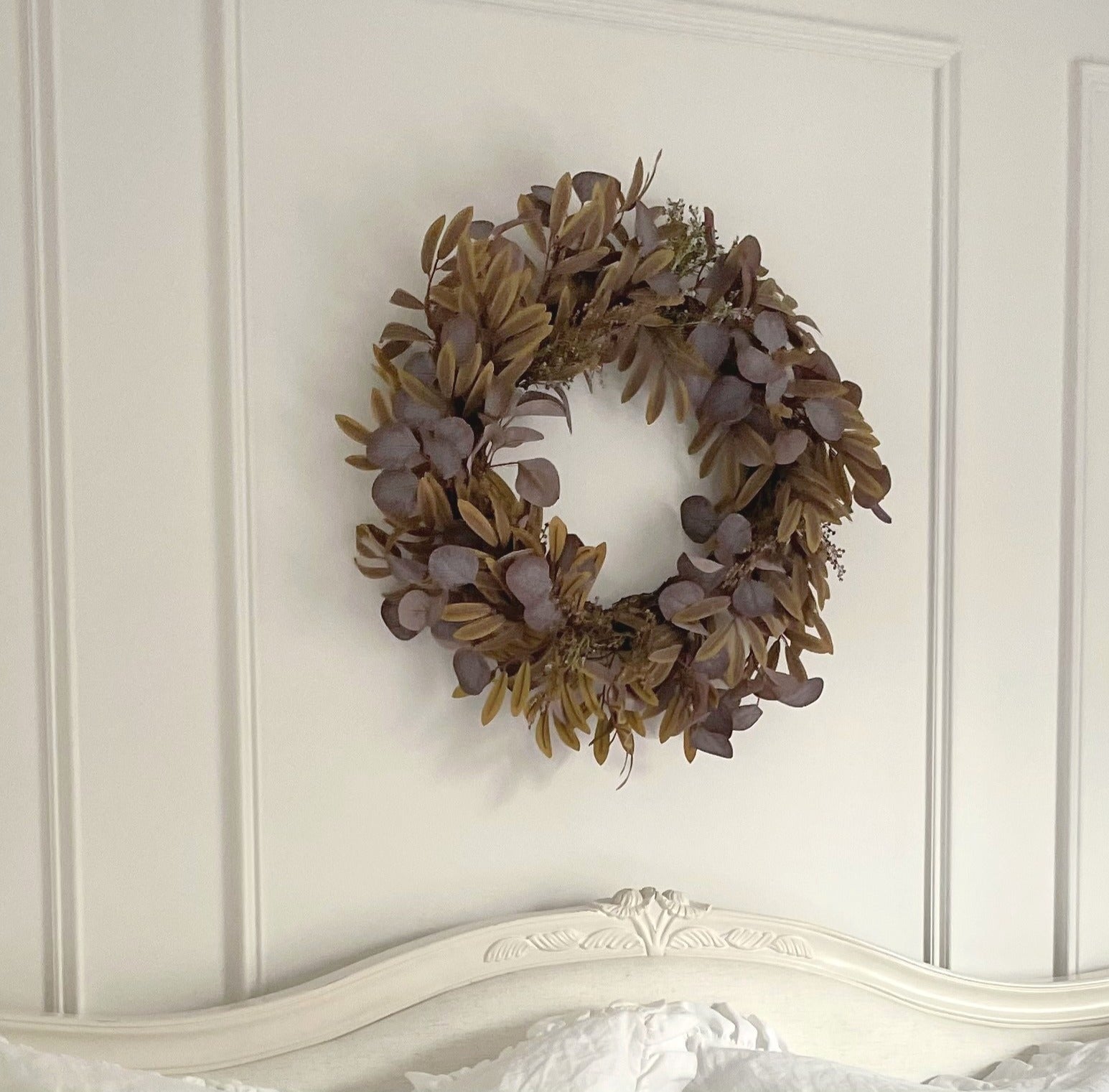 Full autumn foliage wreath with rich fall colours, styled as door or wall décor. Styled on a white panelled wall in a cosy bedroom.