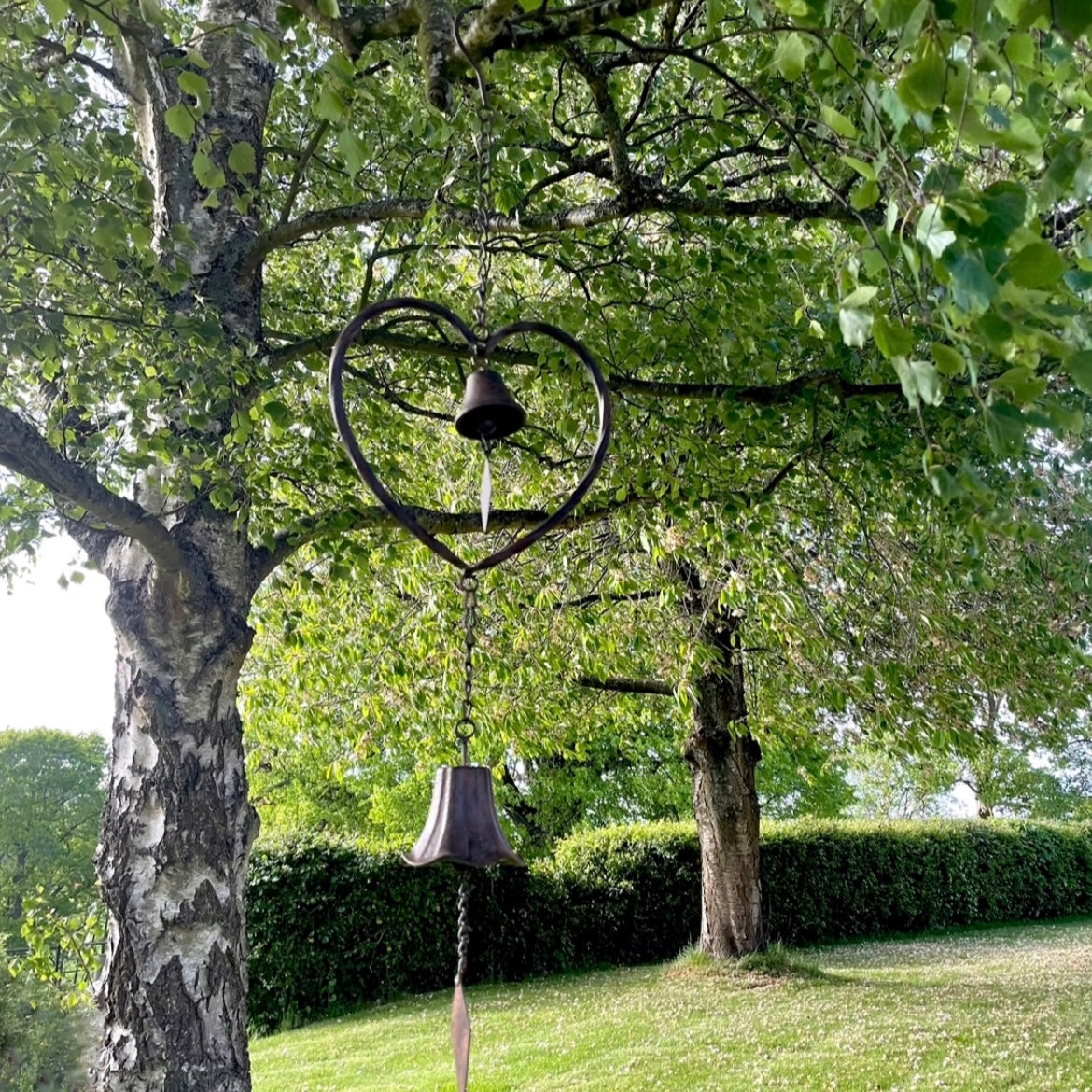 Handmade vintage style heart wind chime with delicate bells, hanging outdoors in a rustic garden setting.
