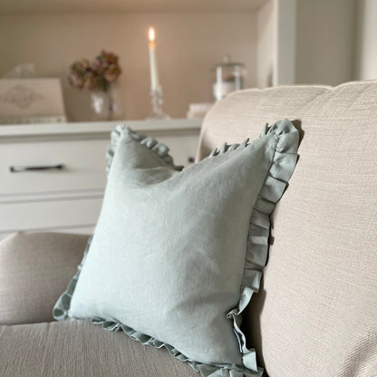 Sage linen cushion with ruffled edge and mother of pearl button detail, styled on a sofa. With shelf and candle in the background.