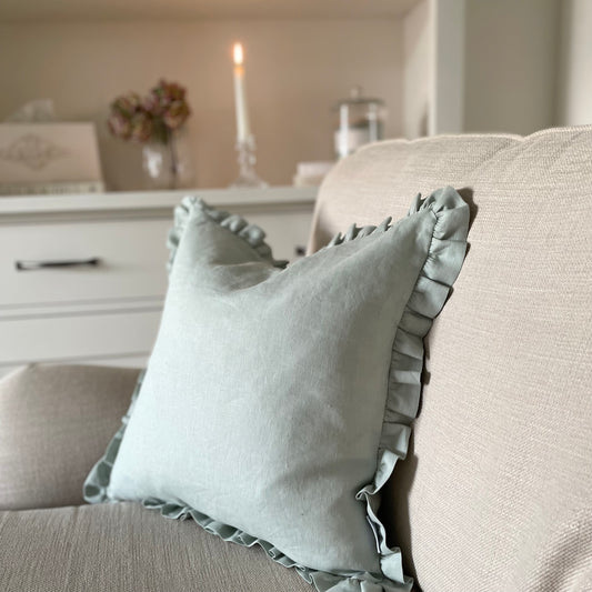 Sage linen cushion with ruffled edge and mother of pearl button detail, styled on a sofa. With shelf and candle in the background.